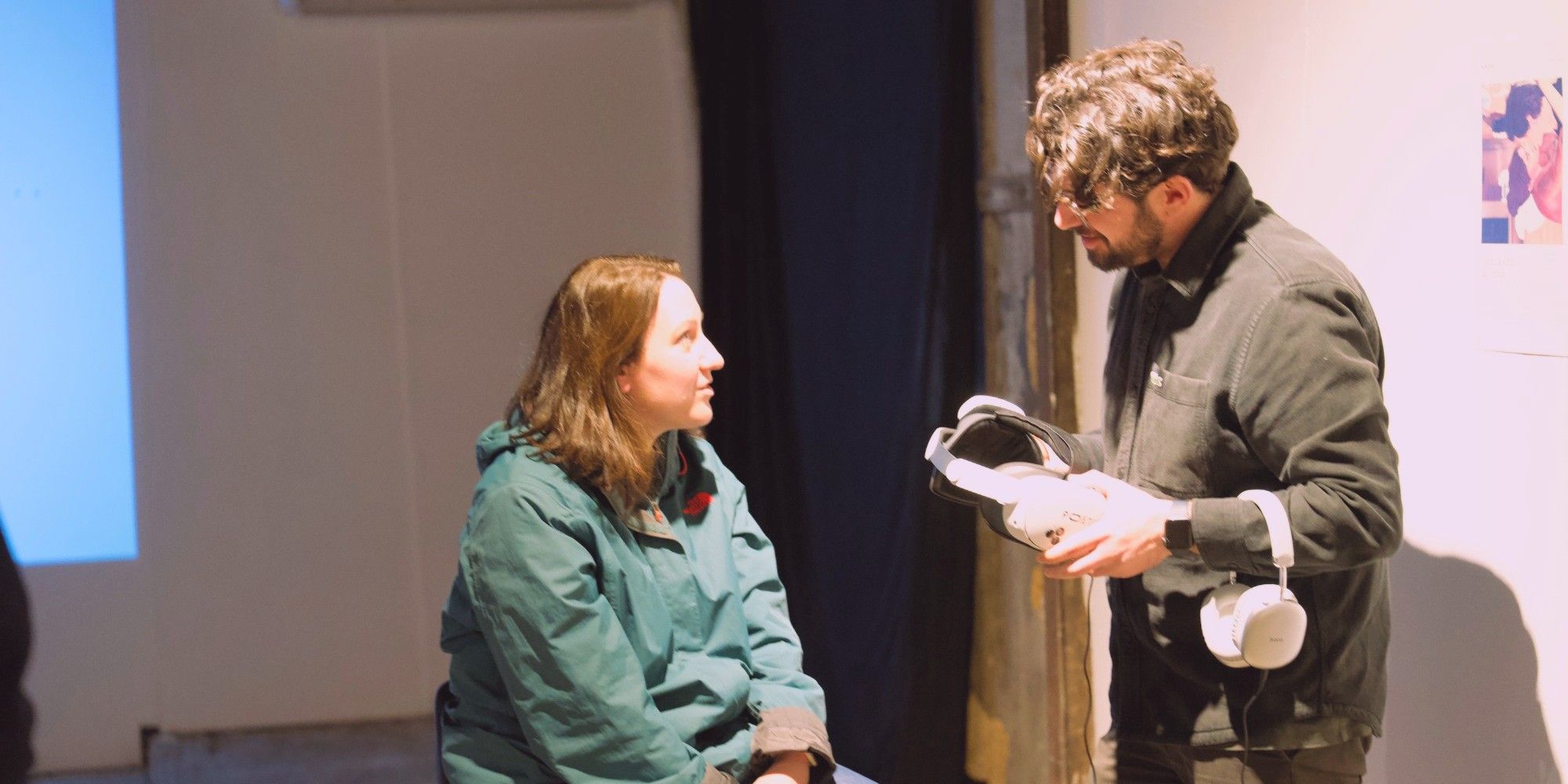 A Poetics operator holding a headset and headphones, talking with a seated visitor before her session.