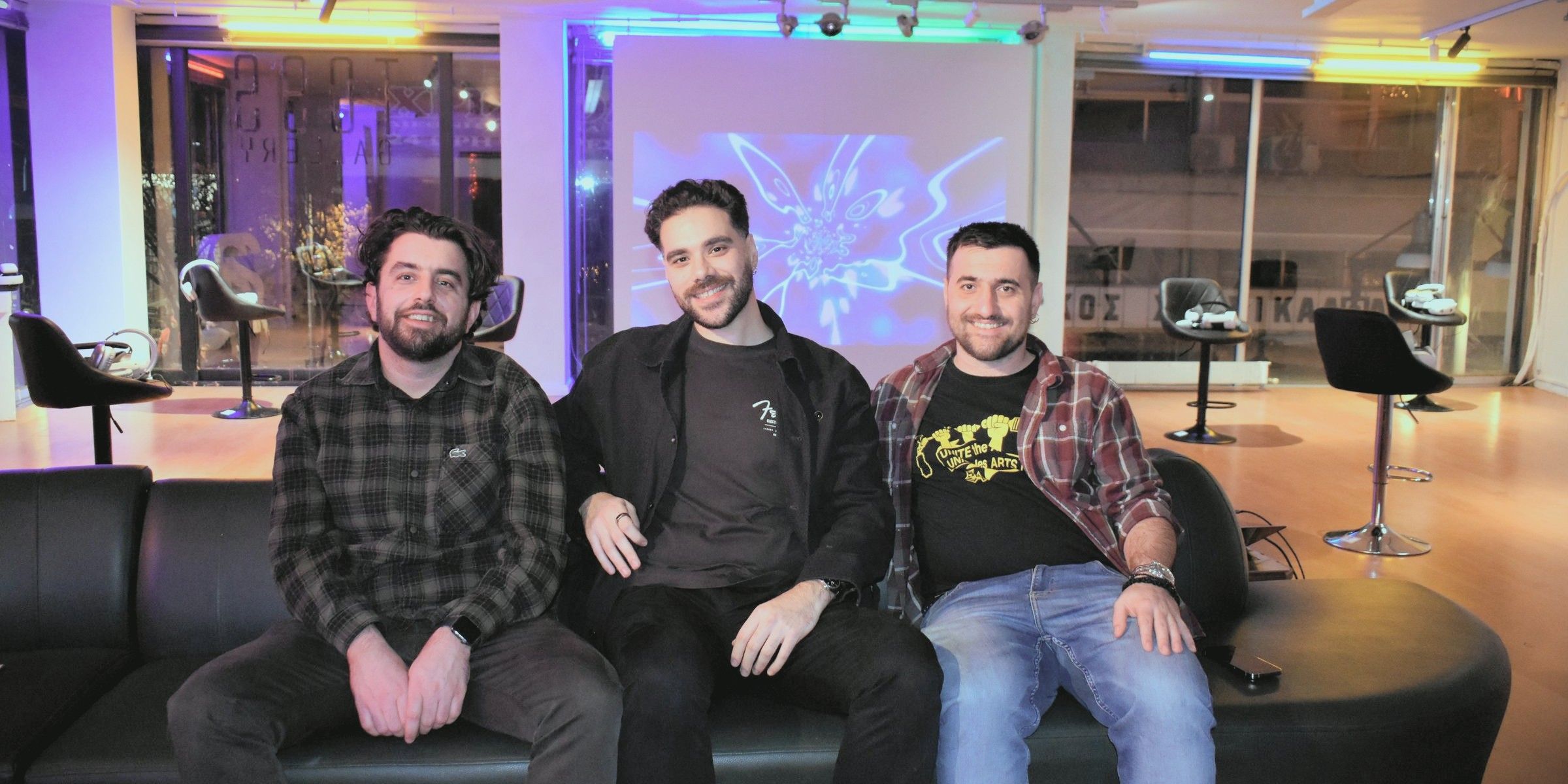 The three founders of Poetics side by side on a black sofa in the pop-up at Toss Gallery, with stools, headsets and a projection behind them.