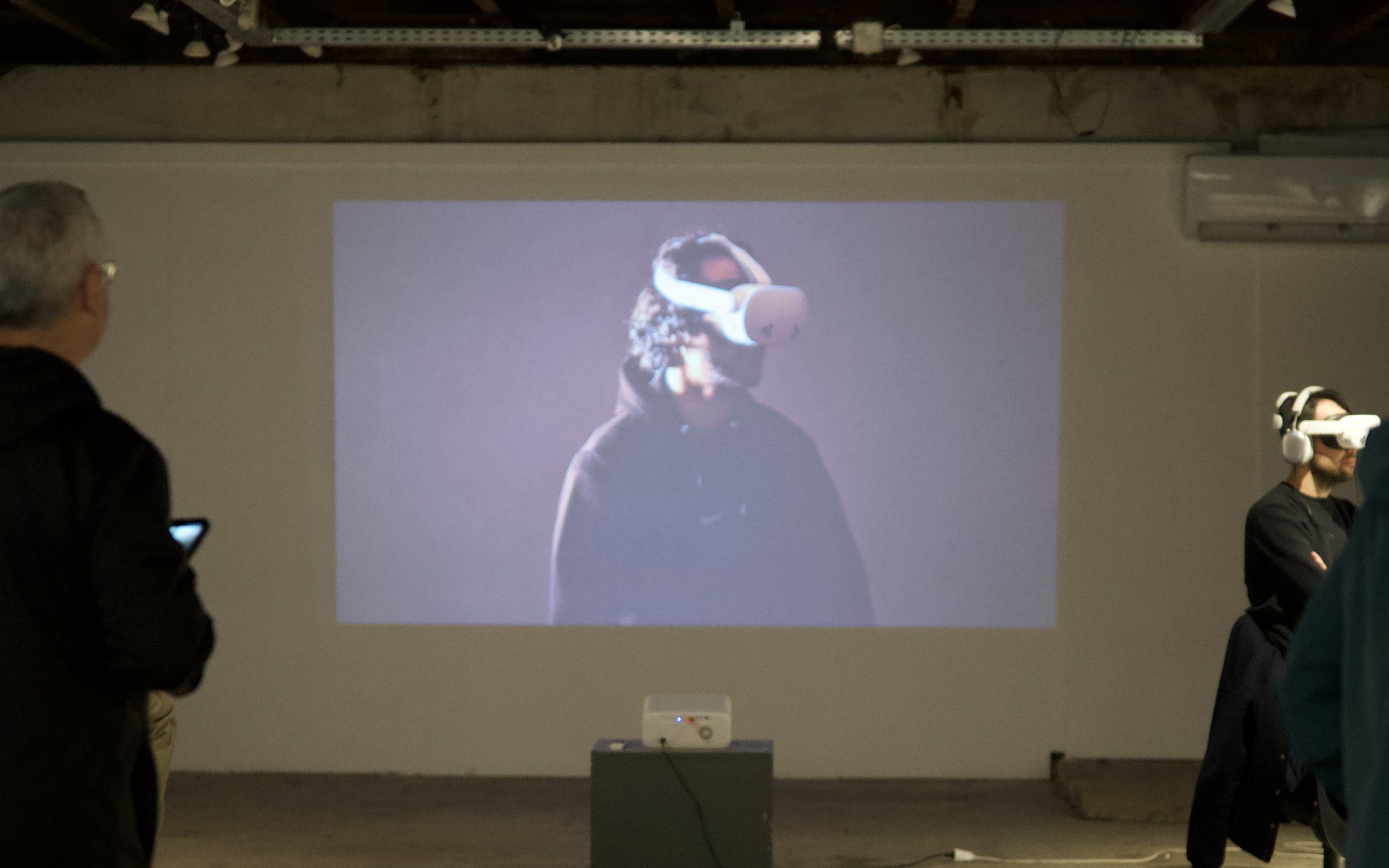 A projected film of a person in a headset in a dark gallery at Technopolis, with a viewer in a headset at the right.