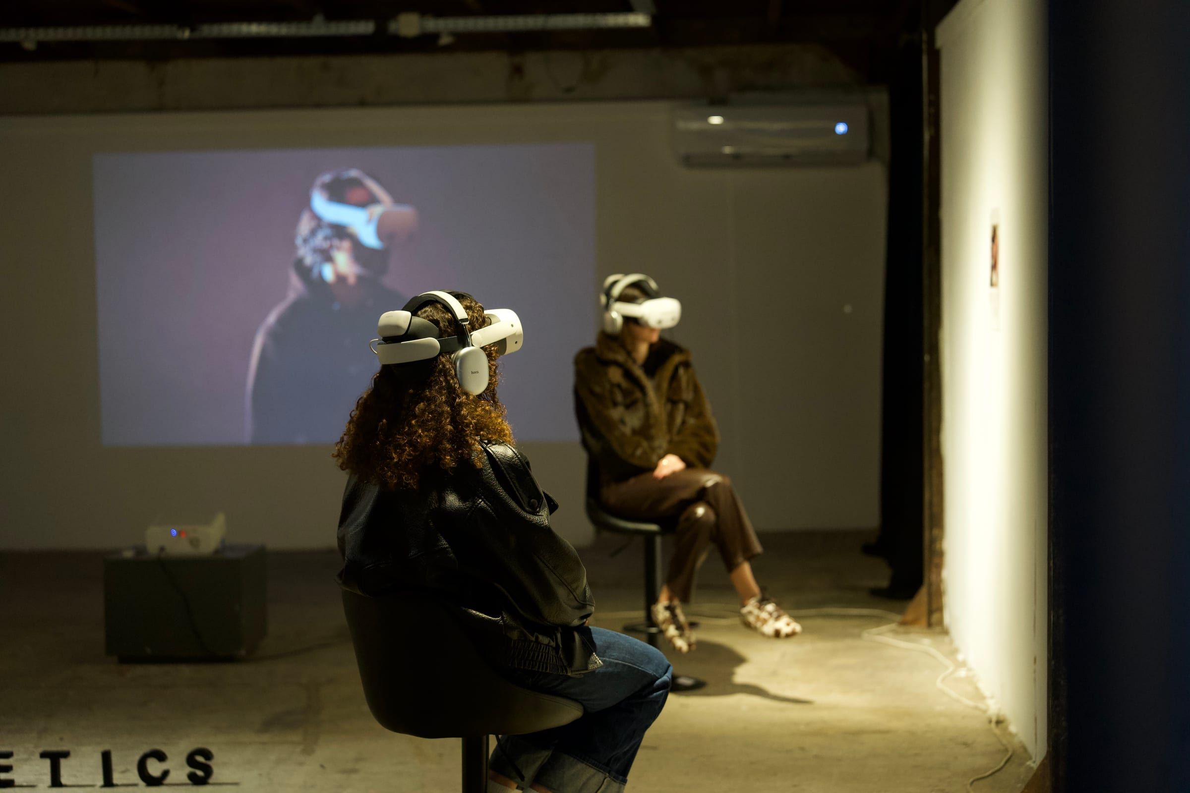 Two visitors in headsets seated in front of a projected film in a gallery.
