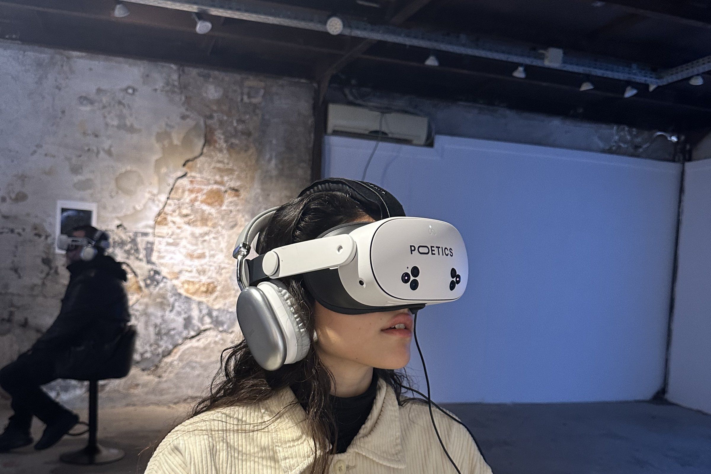 A visitor in a Poetics headset and headphones stands in a gallery with a weathered stone wall, another visitor seated behind her.