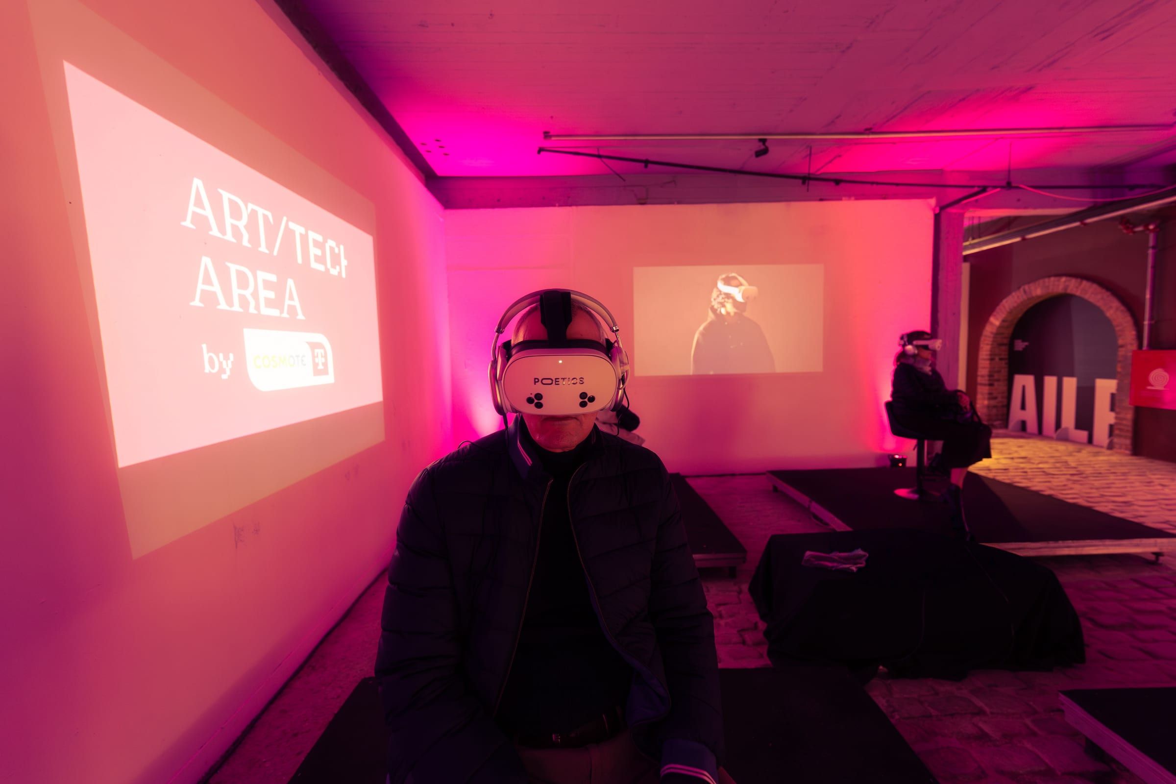 A visitor in a VR headset in a pink-lit room, with a projected Art/Tech Area sign and the festival's AILF arch behind.
