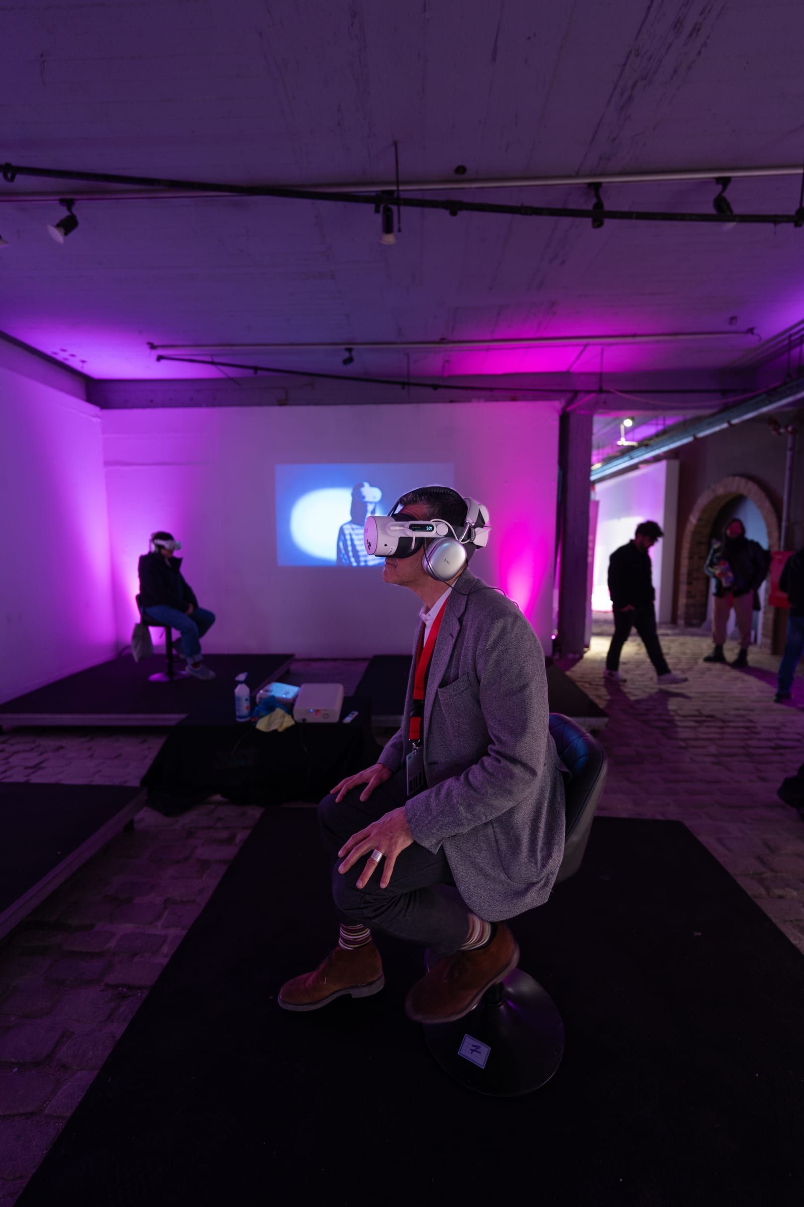 A visitor in a headset in a dark room, with a screen behind and people walking past.