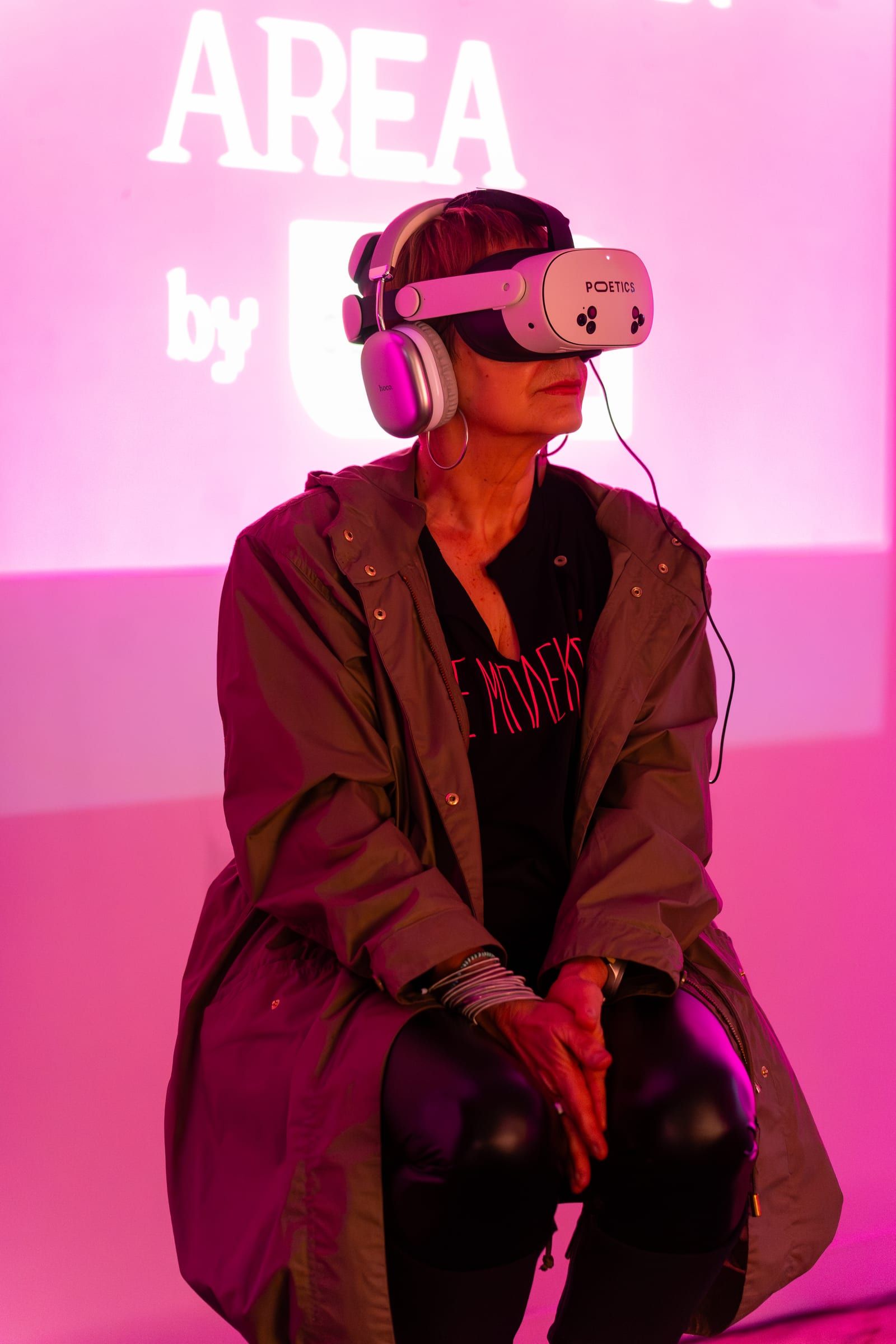 A visitor in a headset and a long green coat, seated in front of an Art/Tech Area backdrop in pink light.