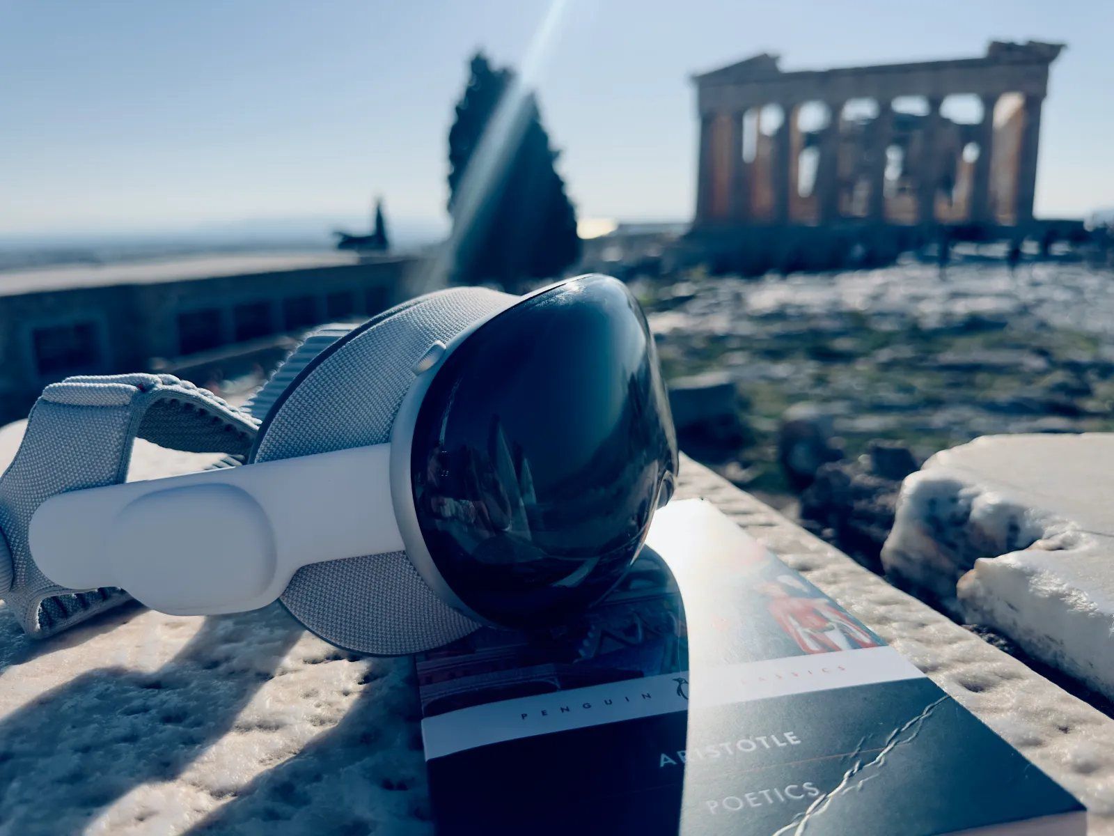 A white VR headset resting on a snowy marble ledge in front of the Parthenon, beside a copy of Aristotle's Poetics.