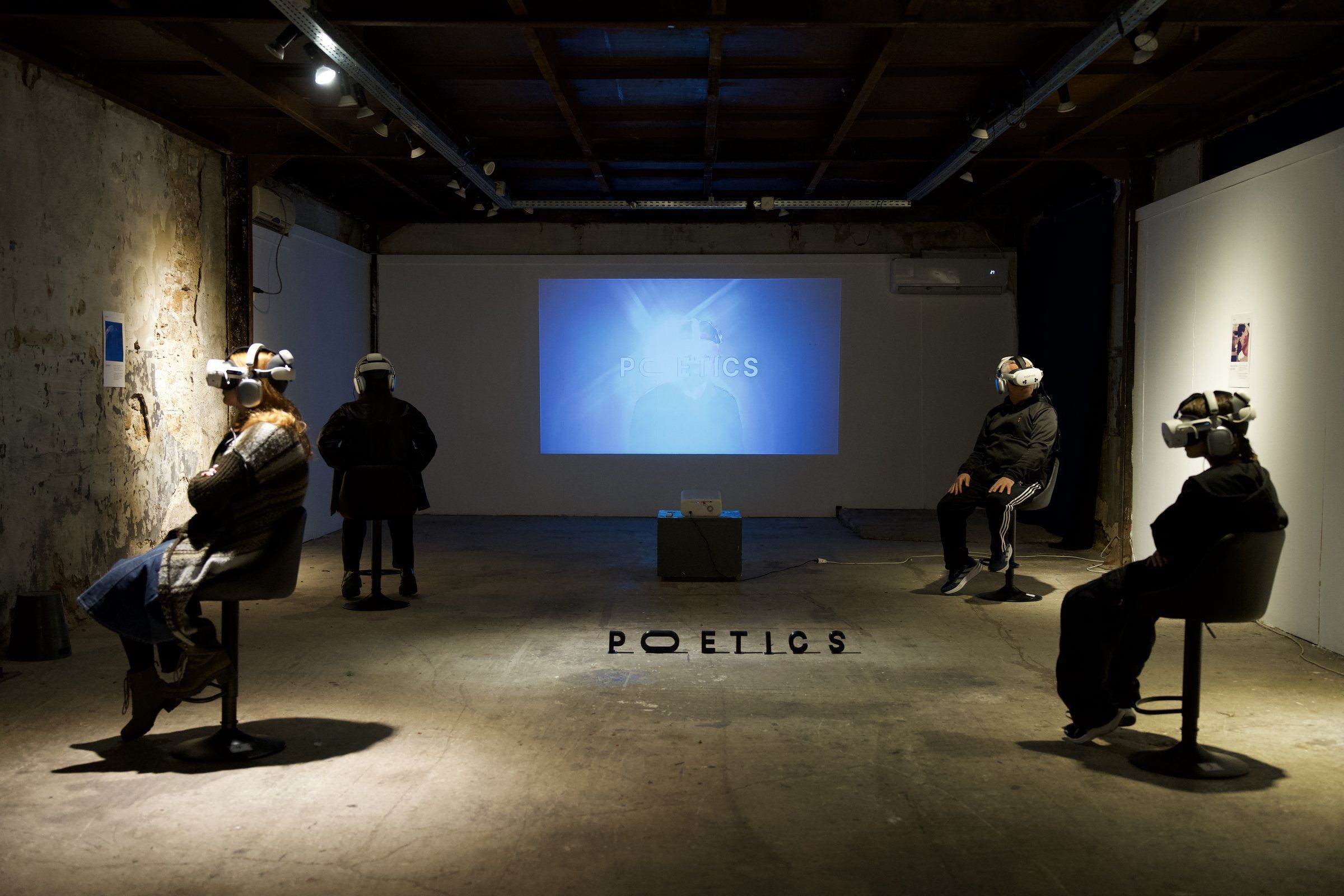 Four visitors on swivel stools wearing headsets face a projected film in a dark gallery, the Poetics name on the floor.