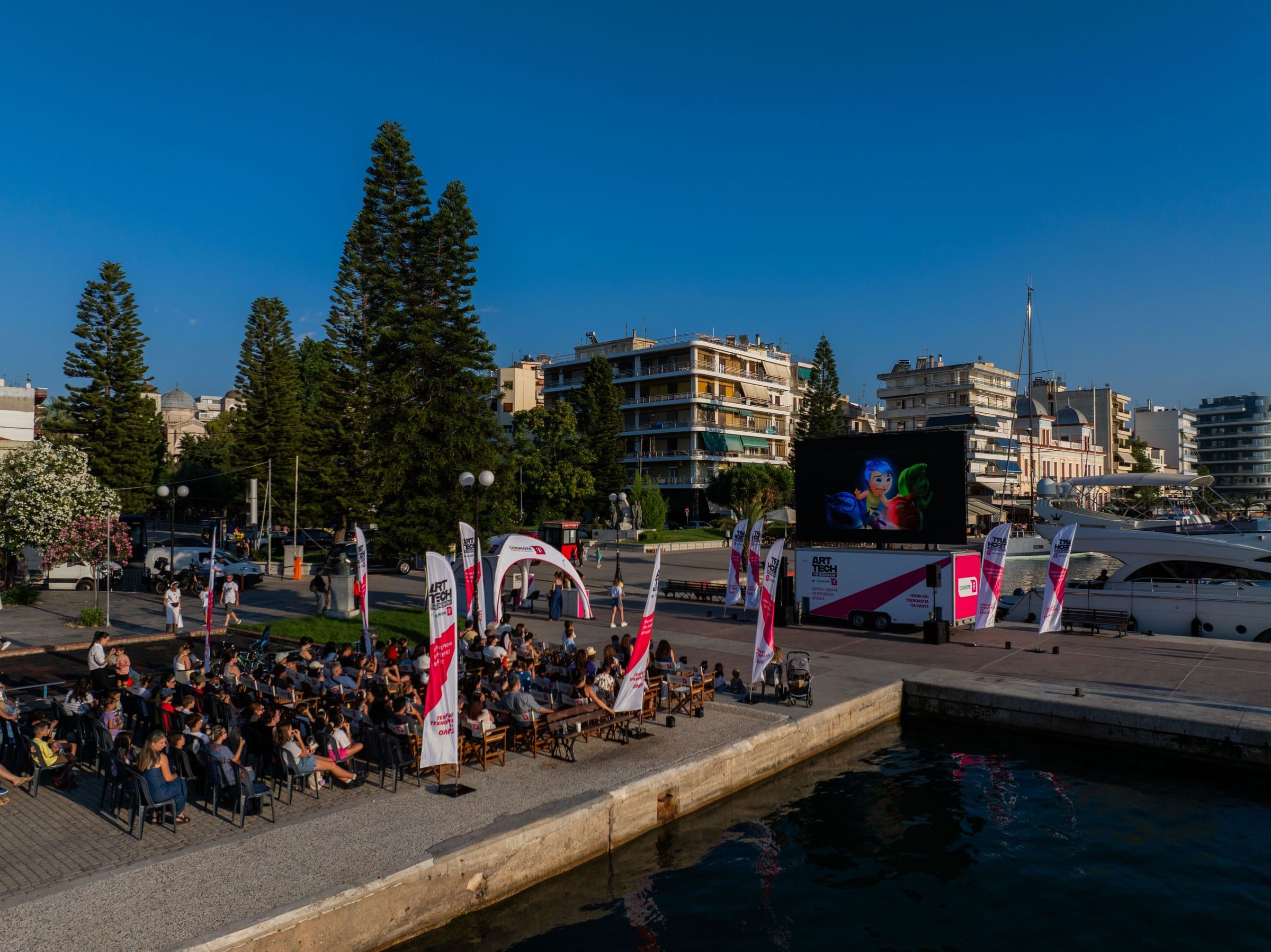 An audience on the Chalkida waterfront watching a big screen at dusk, with ART/TECH flags and tents beside it.