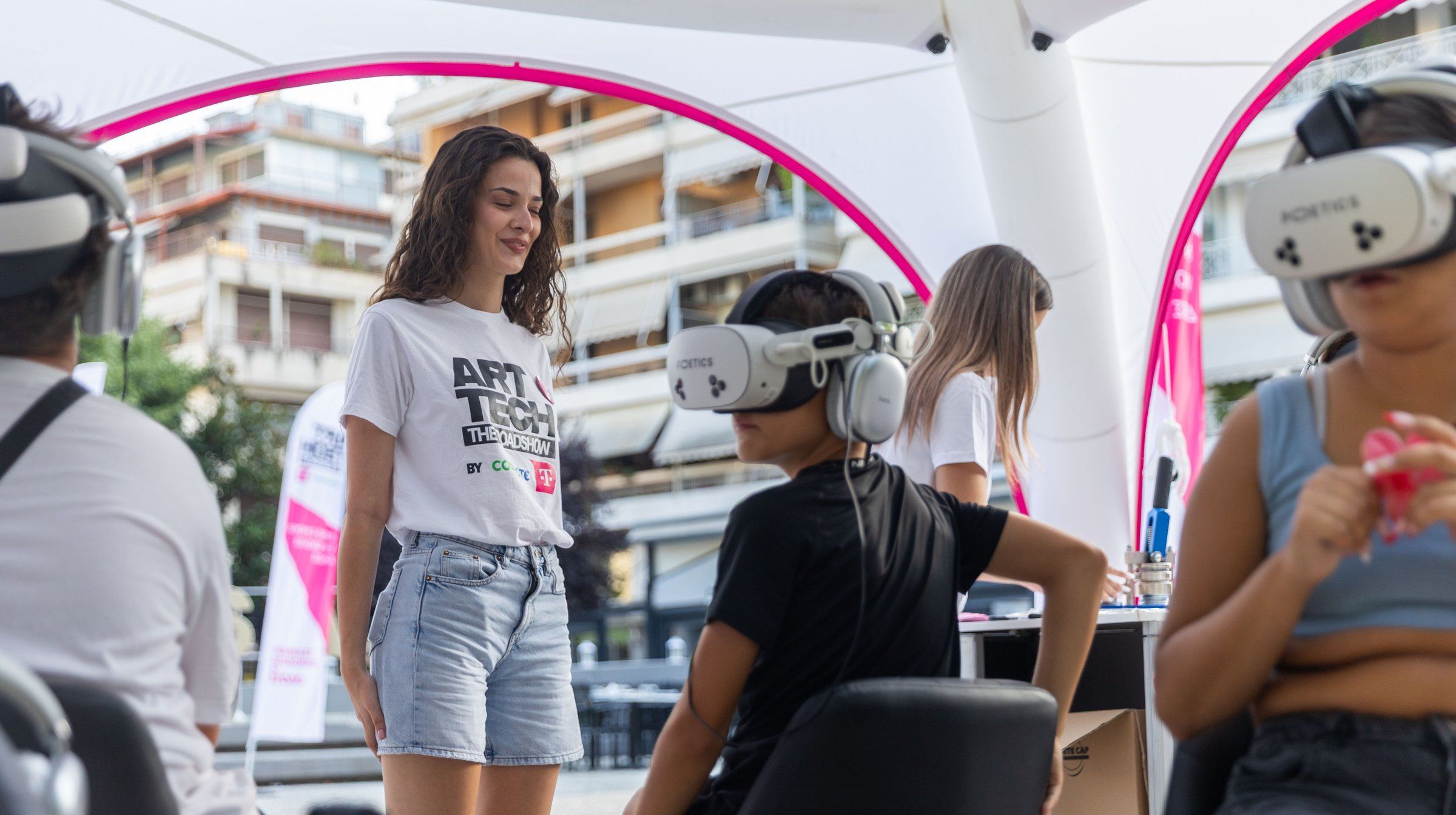 A staff member in an ART/TECH t-shirt stands beside a child in a headset, under the roadshow's white and pink tent.