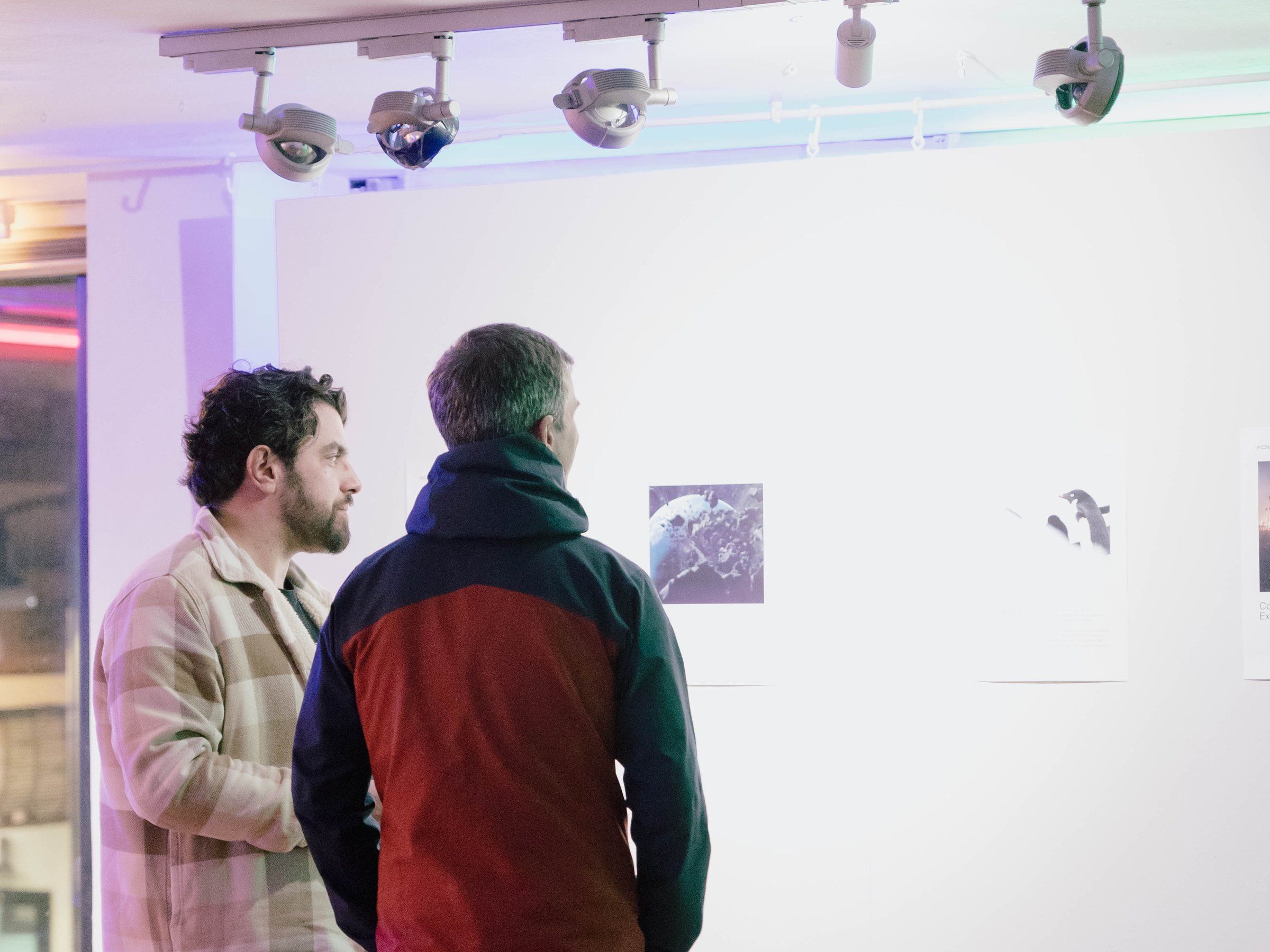 Two visitors study prints on a gallery wall, under spotlights.