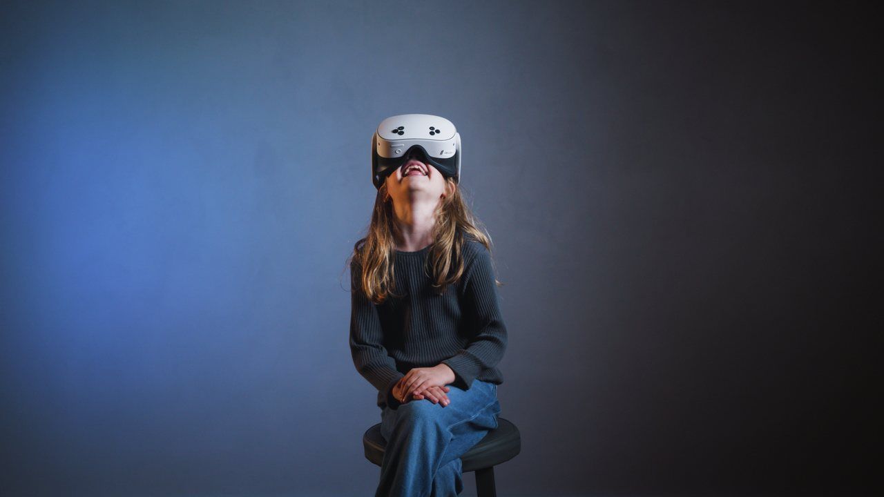 A woman on a stool wearing a headset, smiling and looking up, against a blue wall.