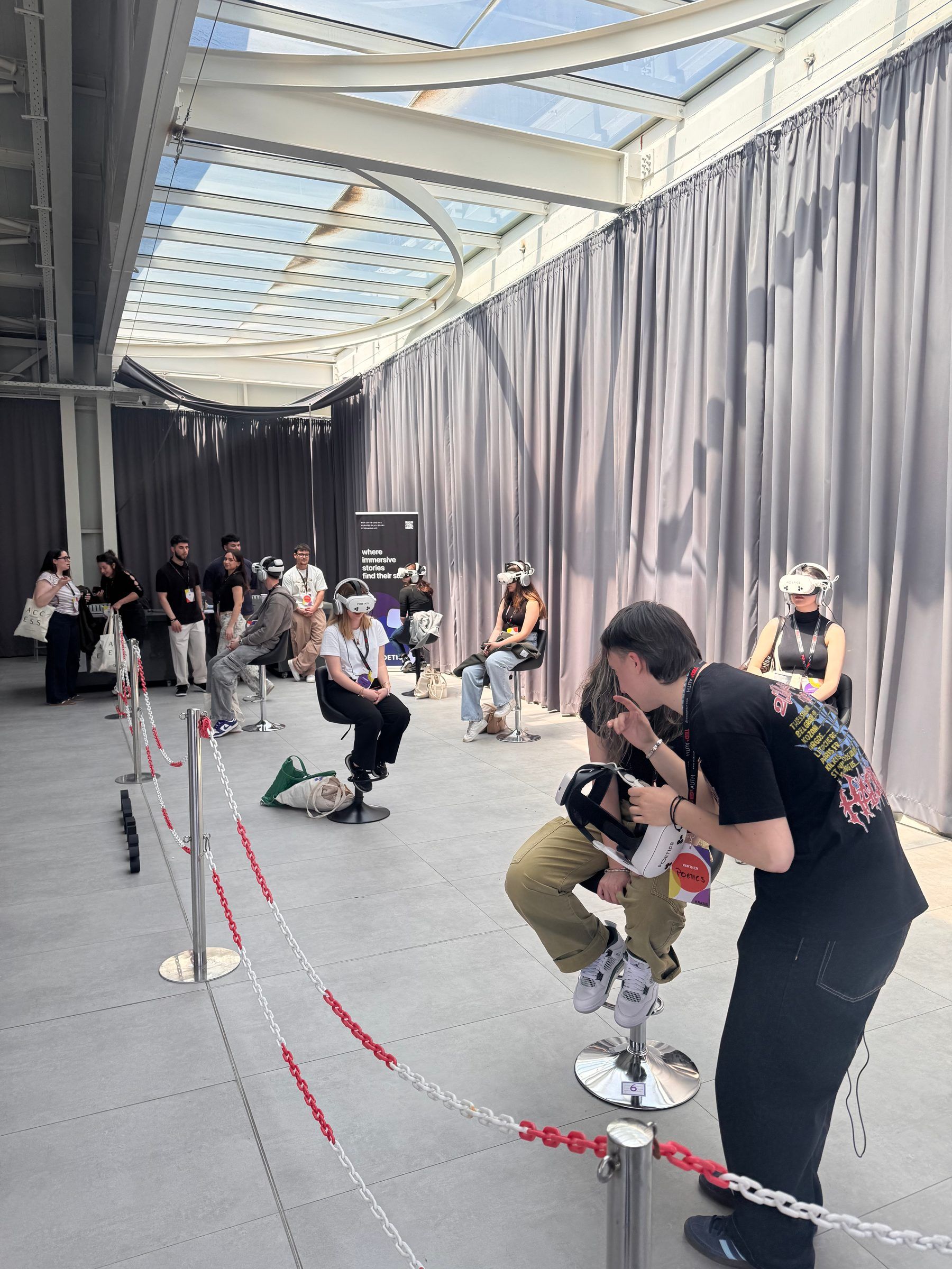 The installation under a glass roof, with viewers in headsets on stools behind a rope line and an operator leaning in to help a visitor with a headset.
