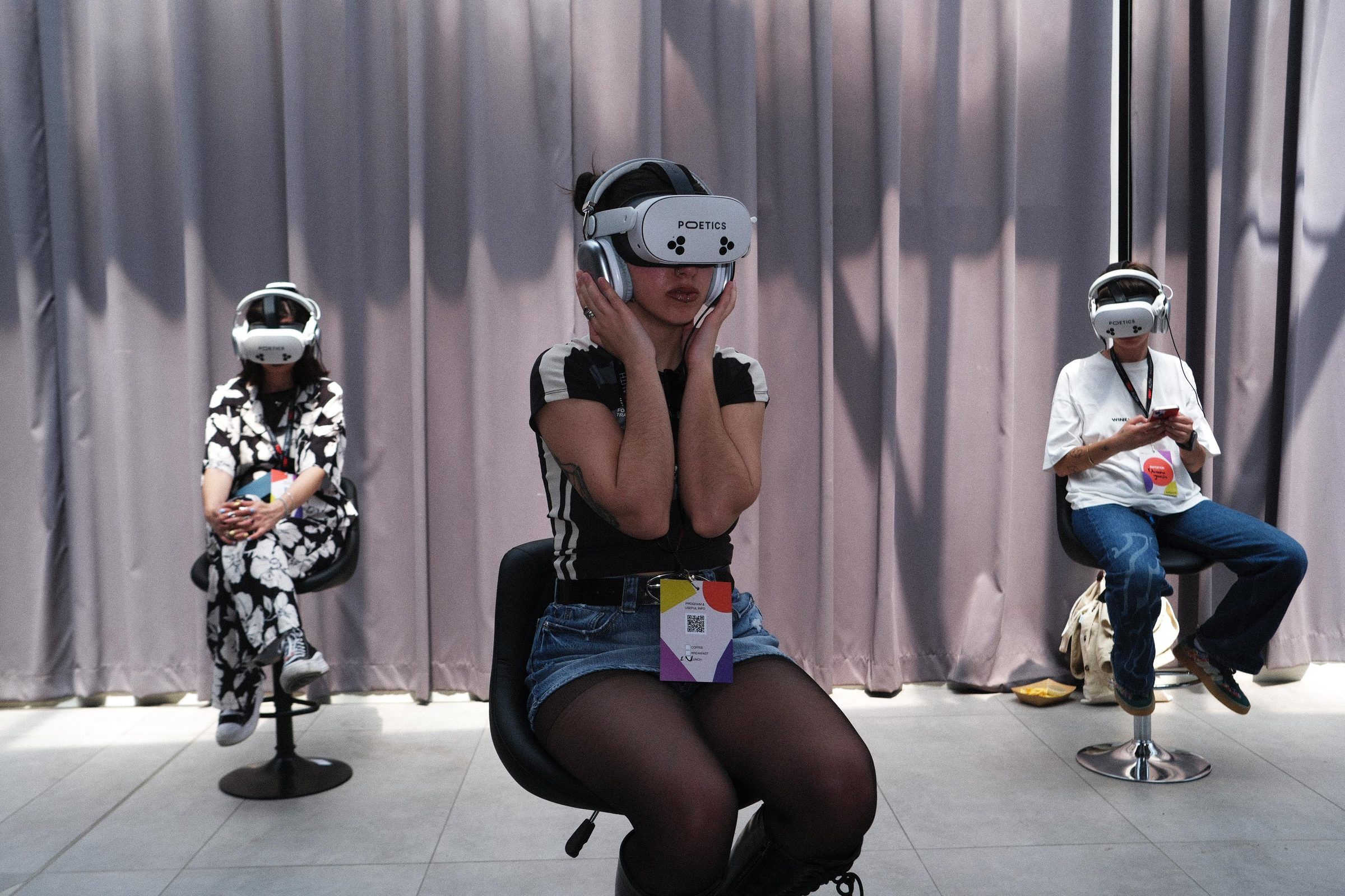 A viewer in a Poetics headset presses the headphones to their ears, with two more viewers in headsets seated against a curtain behind.