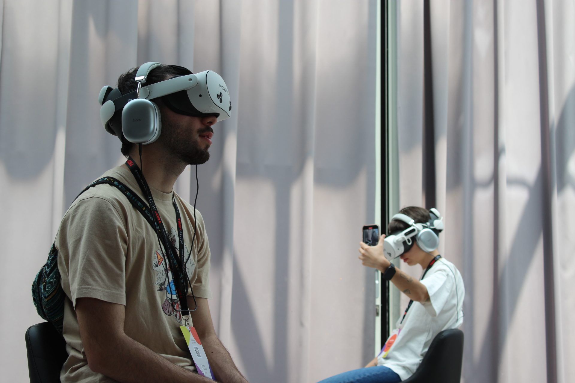 A viewer in a headset and headphones in profile, with another viewer behind taking a photo while still wearing a headset.