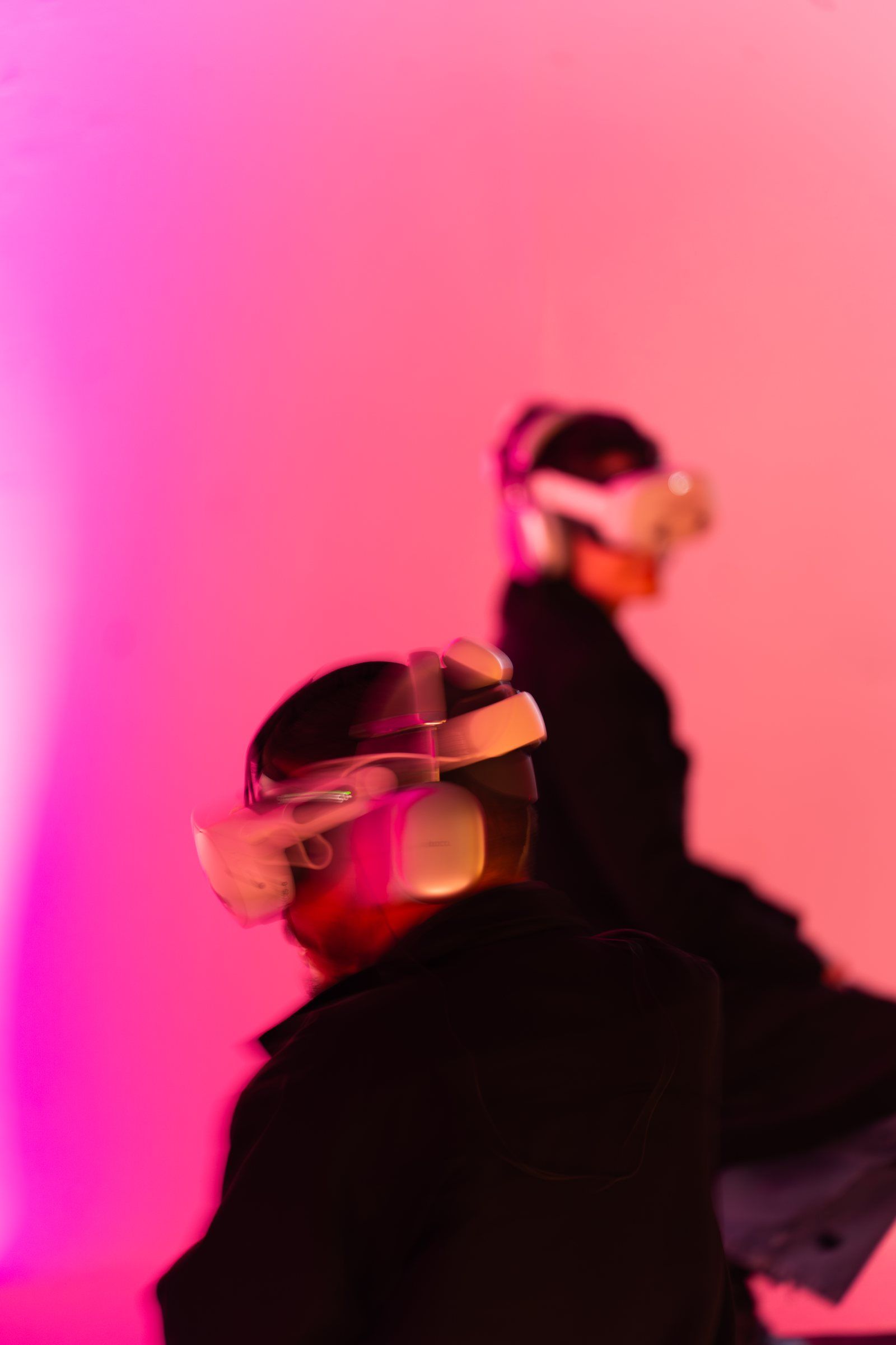 Two visitors in headsets, one seated and one standing, against a wall washed in pink and red light.
